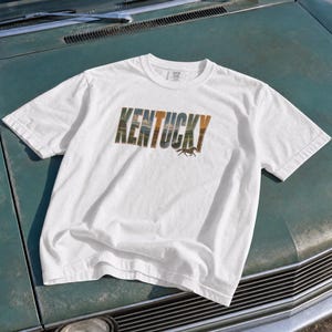May include: A white short-sleeve t-shirt featuring the word KENTUCKY printed in a block font. The letters contain a landscape graphic of rolling hills and trees, with a small silhouette of a running horse positioned below the letter Y.