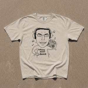 Könnte beinhalten: Ein beiges Kurzarm-T-Shirt mit einer schwarzen Strichzeichnung einer Person, die Kopfhörer trägt und einen Ghettoblaster hält. Der Text "billions must dance" ist unter der Grafik aufgedruckt, begleitet von kleinen Musiknotensymbolen.