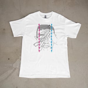 Op de afbeelding: Een wit t-shirt met korte mouwen en een grijze lijntekening van een gezicht. Het ontwerp bevat twee verticale kolommen met kleine menselijke figuren, één in roze en één in blauw, met zwarte stippellijnen die naar het gezicht wijzen.