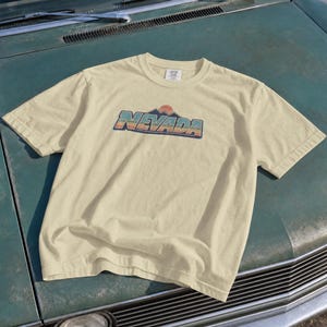Könnte beinhalten: Ein hellgelbes T-Shirt mit kurzen Ärmeln und einem Brustaufdruck, der das Wort NEVADA in einer Retro-Schrift zeigt. Das Design umfasst ein stilisiertes Gebirge und eine Sonne in Orange-, Blau- und Grüntönen. Das Shirt hat einen Rundhalsausschnitt.