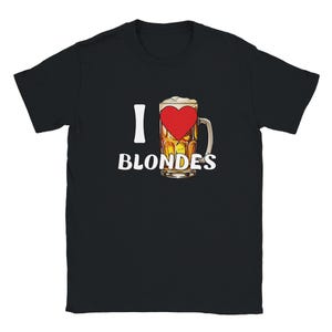Può includere: T-shirt nero con un design grafico. Il design presenta una "I" bianca seguita da un boccale di birra con un cuore rosso al centro e la parola "BLONDES" in testo bianco sotto. Il boccale di birra è pieno di birra.