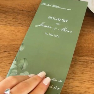Könnte beinhalten: Eine rechteckige, olivgrüne Hochzeitskarte mit weißem Text, der Herzlich Willkommen zur HOCHZEIT von Jessica & Marco 24. Mai 2026 lautet, sowie einer dekorativen Illustration von grünen Blättern in der unteren linken Ecke der Karte.
