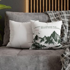 May include: Two white throw pillows on a gray sofa. One pillow features a mountain graphic in dark green with the text "A journey of a thousand miles begins with a single step."