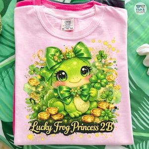 Puede incluir: Camiseta rosa claro Comfort Colors con una princesa rana de dibujos animados. La rana tiene un lazo verde, una corona y tréboles. También se ven monedas de oro y el texto "Lucky Frog Princess 2B". XL-TG-EG.