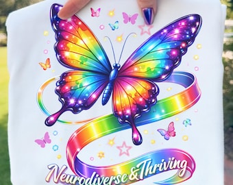 Peut inclure: T-shirt blanc avec un motif de papillon arc-en-ciel vibrant et les mots "Neurodiverse & Thriving" sur un ruban arc-en-ciel. Le motif comprend des papillons et des étoiles supplémentaires.