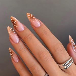 Può includere: Unghie a mandorla con base color carne, punte marroni e puntini bianchi. Le dita sono adornate con diversi anelli color argento, tra cui un anello con una pietra centrale trasparente e una fascia spessa e liscia sull'indice.