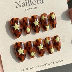 Könnte beinhalten: Ein Set aus zehn mandelförmigen Press-on-Nägeln mit glänzendem braun-schwarzem Schildpattmuster und einem hellgelben Sternendesign auf jedem Nagel. Die Nägel sind auf einer weißen Karte mit der Aufschrift Naillora press on nail handmade reusable customisable befestigt.