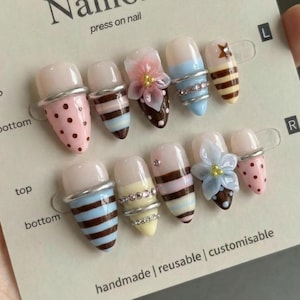 Op de afbeelding: Een set persnagels met diverse ontwerpen, waaronder roze stippen, bruine strepen en bloemaccenten. De nagels worden gepresenteerd op een kaart met de tekst "Naillora press on nail" en "handmade | reusable | customisable".