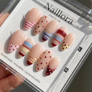 Op de afbeelding: Een set van tien amandelvormige press-on nagels in een doorzichtig plastic doosje met het label Naillora. De nagels hebben een nude basis met diverse ontwerpen, waaronder rode en blauwe horizontale strepen en rode of bruine stippen.