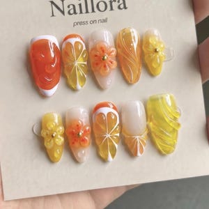 May include: A set of press-on nails with a citrus fruit theme. The nails feature orange, yellow, and white designs, including orange slices and flower accents. The brand name "Naillora" is printed on the card.