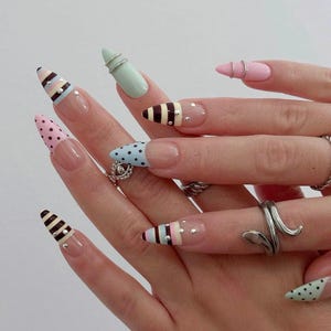 Puede incluir: Una mano con uñas largas en forma de almendra, cada una pintada con un diseño único. Las uñas presentan patrones como lunares, rayas y colores pastel, acentuados con pequeños pedrería y anillos plateados. La estética general es una mezcla de arte de uñas juguetón y elegante.