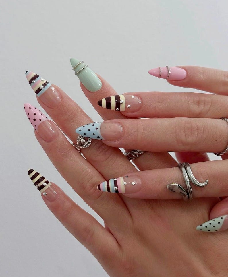 May include: A hand with long, almond-shaped nails, each painted with a unique design. The nails feature patterns like polka dots, stripes, and pastel colors, accented with small rhinestones and silver rings. The overall aesthetic is a blend of playful and elegant nail art.