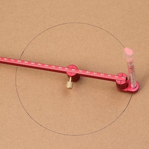 May include: A red compass tool with a ruler and a clear plastic tube is used to draw a circle on a brown surface. The compass has measurements in millimeters and a brass-colored adjustment knob.