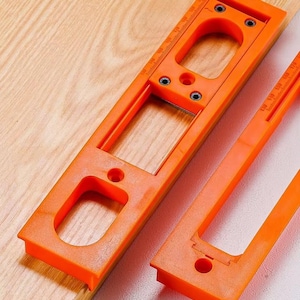 Könnte beinhalten: Zwei leuchtend orangefarbene Holzbearbeitungsvorrichtungen mit Maßstäben. Eine Vorrichtung ist rechteckig mit einem großen Ausschnitt und Schraubenlöchern. Die andere ist kleiner, mit einem ähnlichen Design. Beide liegen auf einer hellen Holzoberfläche.