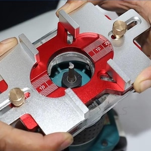 May include: A metal router base with red and silver components. The base has a circular opening with a central spindle and measurement markings. The tool is held by hands, showcasing its design for woodworking and precision tasks.
