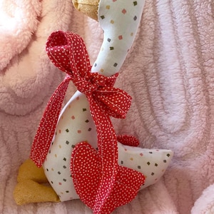 May include: A stuffed fabric goose toy featuring a white body with a small multicoloured diamond pattern, a gold textured beak and feet, and a bright red ribbon with white polka dots tied around its neck with a matching red heart-shaped wing.