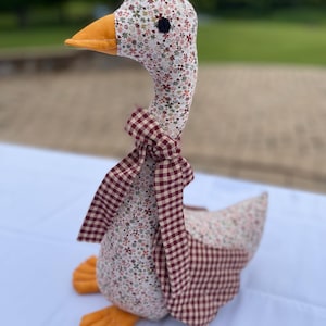 May include: A fabric goose toy with a floral patterned head and body, and a burgundy gingham bow. The goose has an orange beak and feet. The background is blurred, suggesting an outdoor setting.