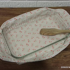 Könnte beinhalten: Eine transparente Glasbackform und ein Holzspatel liegen in einem floralen Stoffbezug. Der Stoff ist hellgrau mit einem Muster aus kleinen roten Blumen und grünen Blättern. Die Form liegt auf einer Holzoberfläche.