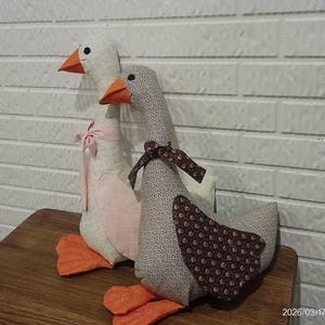 May include: Two handmade goose figurines. One is white with a pink bow, the other is gray with a brown patterned wing. Both have orange beaks and feet. They are sitting on a wooden surface.