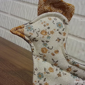 May include: A fabric chicken-shaped decorative item. The chicken has a cream-colored body with a floral pattern in shades of orange, blue, and brown. The beak and comb are a solid brown color. The item is likely handmade and placed on a wooden surface.