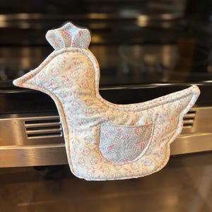 以下が含まれることがあります： ピンク、青、クリーム色の花柄の、アヒルの形をしたオーブンミトン。ミトンには小さなポケットと、上部に王冠のようなディテールがあります。キルティング生地で作られており、手を熱から保護するように設計されています。