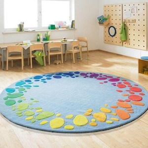 Round kids play rug with colorful rainbow pebble pattern on soft blue background placed in a bright playroom with wooden floor and small chairs. Modern nursery and classroom rug designed for toddlers and children. Soft circle play mat ideal for playroom, daycare, preschool or kids bedroom. Available in multiple round sizes, this colorful rug adds comfort, safety and fun learning space for kids in homes worldwide.