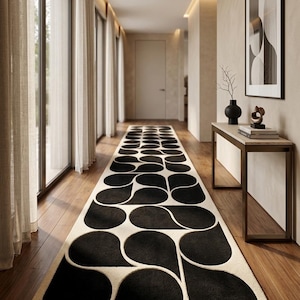 May include: A long, rectangular rug with a repeating pattern of black teardrop shapes on a cream background. The rug is placed in a hallway with wooden flooring and large windows with sheer curtains. The hallway also features a console table with decor and a framed artwork.