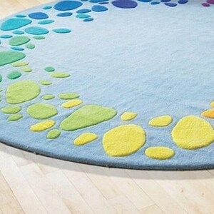 Round kids play rug with colorful rainbow pebble pattern on soft blue background placed in a bright playroom with wooden floor and small chairs. Modern nursery and classroom rug designed for toddlers and children. Soft circle play mat ideal for playroom, daycare, preschool or kids bedroom. Available in multiple round sizes, this colorful rug adds comfort, safety and fun learning space for kids in homes worldwide.