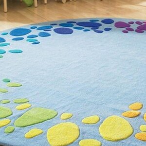 Round kids play rug with colorful rainbow pebble pattern on soft blue background placed in a bright playroom with wooden floor and small chairs. Modern nursery and classroom rug designed for toddlers and children. Soft circle play mat ideal for playroom, daycare, preschool or kids bedroom. Available in multiple round sizes, this colorful rug adds comfort, safety and fun learning space for kids in homes worldwide.