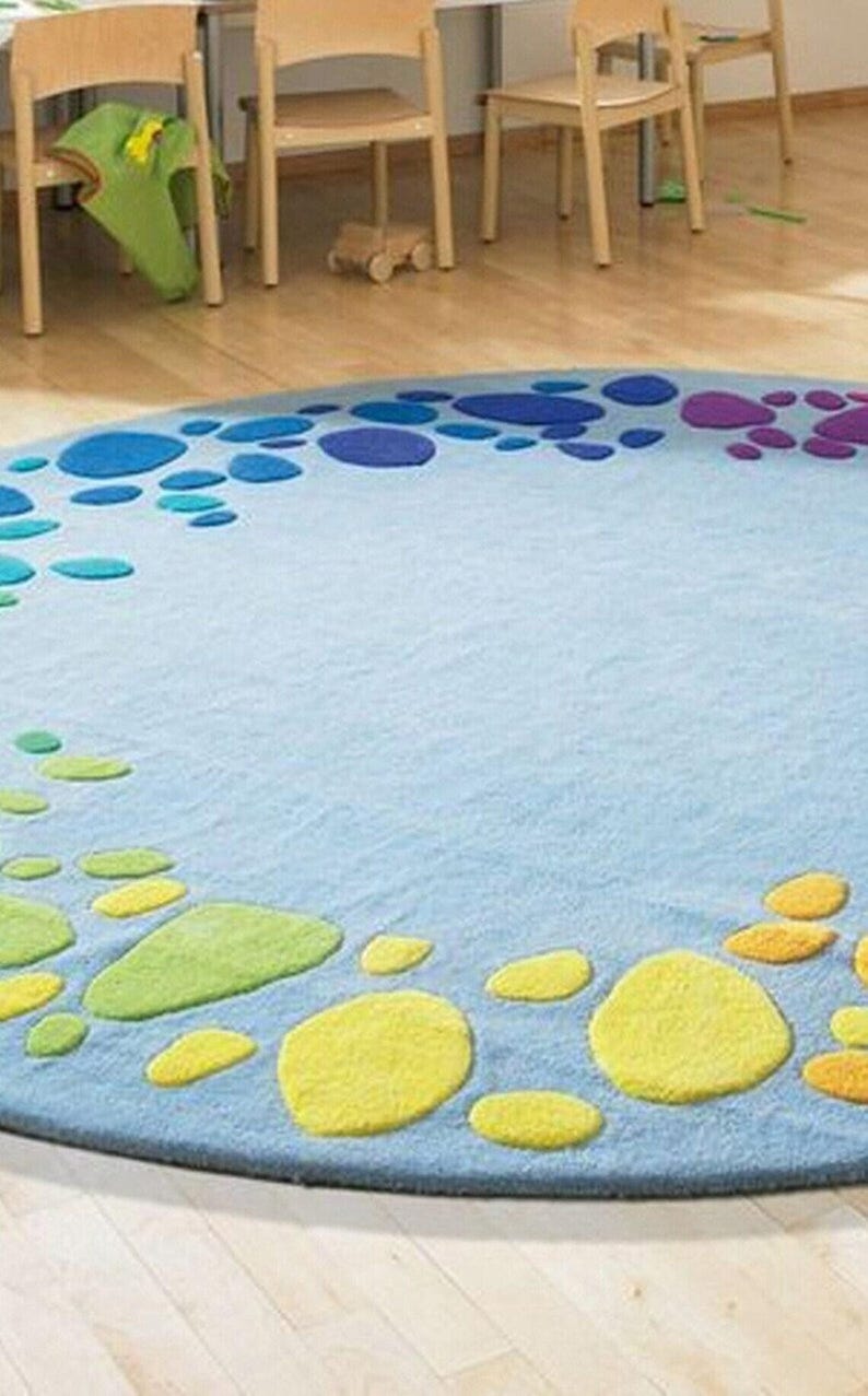 Round kids play rug with colorful rainbow pebble pattern on soft blue background placed in a bright playroom with wooden floor and small chairs. Modern nursery and classroom rug designed for toddlers and children. Soft circle play mat ideal for playroom, daycare, preschool or kids bedroom. Available in multiple round sizes, this colorful rug adds comfort, safety and fun learning space for kids in homes worldwide.