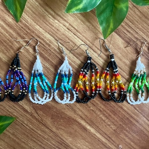 Può includere: Una collezione di orecchini di perline fatti a mano in vari colori, tra cui blu, viola, bianco, turchese, giallo, rosso e verde. Gli orecchini hanno una forma a goccia e sono esposti su una superficie di legno.