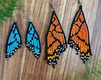May include: Four pairs of beaded earrings in a butterfly wing design. Two pairs are turquoise and black, and two are orange, yellow, and black. The earrings have a beaded fringe at the bottom and are displayed on a wooden surface.