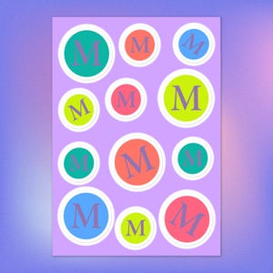 May include: A sheet of stickers with a repeating pattern of the letter "M" inside colorful circles. The circles are in various colors, including green, orange, blue, and pink, with a white border, set against a lavender background.