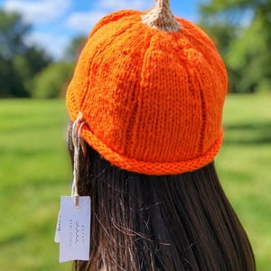 May include: A bright orange knitted beanie shaped like a pumpkin with a small tan stem at the top. The hat features a ribbed texture and a rolled brim. A white rectangular tag with the text NANI STUDIO is attached to the side with a string.