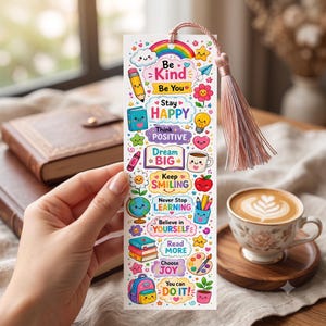 May include: A rectangular paper bookmark featuring a light pink tassel and colourful illustrations of a rainbow, pencil, books, globe, and coffee cup. The text reads: Be Kind, Be You, Stay Happy, Think Positive, Dream Big, Keep Smiling, Never Stop Learning, Believe in Yourself, Read More, Choose Joy, You can DO IT.