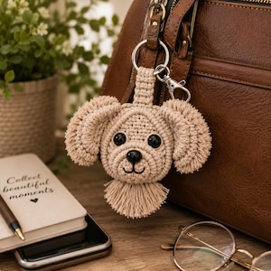 May include: A beige macrame dog keychain featuring a round head with textured floppy ears, black bead eyes, a black nose, and a small fringe at the bottom. The keychain is attached to a silver metal lobster clasp and a circular ring connector.