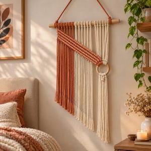 Può includere: Un arazzo in macramè con un bastone in legno, cordoncini di cotone color crema e terracotta e un anello centrale in legno. Il design presenta una sezione diagonale di cordoncini color terracotta annodati sopra cordoncini verticali color crema.