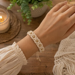 Op de afbeelding: Een crèmekleurige macramé armband met een herhalend ruitvormig knooppatroon met kleine, gepolijste goudkleurige metalen kralen in het midden van elke ruit. De armband heeft verstelbare koorduiteinden met kleine kralen aan de uiteinden.