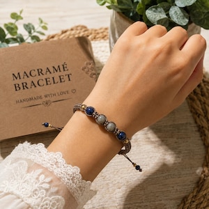 Op de afbeelding: Een macramé armband met twee centrale grijze stenen kralen geflankeerd door twee donkerblauwe lapis lazuli kralen en kleine goudkleurige metalen accenten, geweven in een bruin koord met verstelbare trekkoorden en kleine eindkralen.