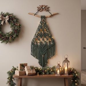 Könnte beinhalten: Ein dunkelgrüner Makramee-Wandbehang in Form eines Weihnachtsbaums, der an einem Holzast mit geschnitztem Vogelornament hängt. Das Stück ist mit komplexen Knoten gewebt und mit kleinen Sternanhängern sowie warmweißen Lichterketten verziert.