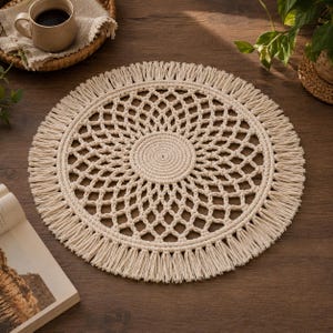 May include: A round, off-white macrame placemat featuring a central spiral knot pattern surrounded by an intricate, open-work diamond lattice design. The circular mat is finished with a dense, uniform fringe border made of twisted cotton cord.