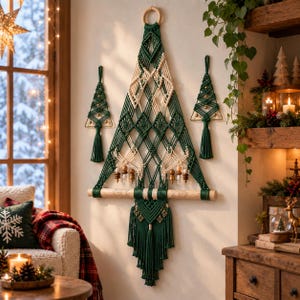 May include: A large macrame wall hanging shaped like a Christmas tree, crafted from dark green and cream-coloured cord. It features intricate diamond patterns, wooden beads, and a horizontal wooden dowel at the base, flanked by two smaller matching tree ornaments.