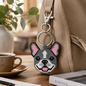 Peut inclure: Un porte-clés en perles représentant un bouledogue français aux tons gris, blancs et noirs avec des oreilles intérieures roses. La breloque est fixée à un mousqueton et un anneau en métal argenté, suspendue à un sac en tissu beige.