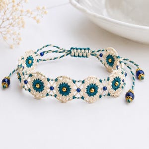 May include: A cream-coloured crochet bracelet with teal and gold detailing. The bracelet features a floral pattern with blue beads and teal and cream-coloured cords. The bracelet is adjustable.