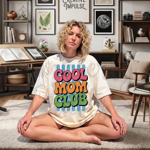 Puede incluir: Camiseta color crema con el texto "COOL MOM CLUB" en una fuente retro. El texto es rosa, naranja y verde, con rayas verticales azules encima. La camiseta es de corte holgado.