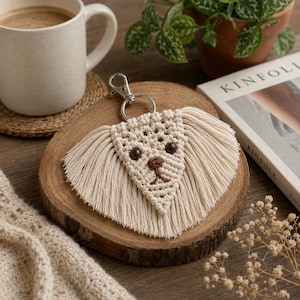 Peut inclure: Un porte-clés en macramé couleur crème en forme de visage de chien, avec de longues oreilles à franges, deux yeux en perles marron foncé et un nez ainsi qu'une bouche brodés en marron. Il comporte un mousqueton et un anneau en métal argenté.