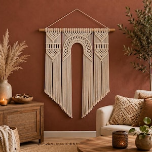 May include: A large off-white macramé wall hanging suspended from a horizontal wooden dowel with a string. The textile art features intricate knotted patterns, including a central arch design flanked by two geometric panels with long fringe.