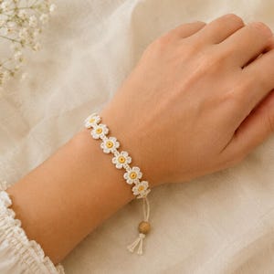 Puede incluir: Una pulsera blanca de ganchillo con diseños de flores de margarita con centros amarillos. La pulsera se lleva en la muñeca y tiene un cierre con una cuenta de madera y borlas. El fondo es de un color suave y neutro.