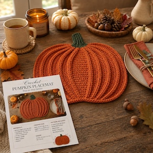 May include: A crocheted pumpkin-shaped placemat in orange with a dark green stem, featuring a ribbed texture. Beside it lies a printed instruction sheet titled Crochet PUMPKIN PLACEMAT, detailing materials, finished size of 15-16 inches, and gauge.