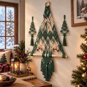Puede incluir: Un gran tapiz de macramé con forma de árbol de Navidad, elaborado con cuerda de color verde oscuro y crema. Presenta intrincados patrones de nudos en forma de diamante, cuentas de madera y un soporte de madera horizontal, junto a dos adornos a juego.